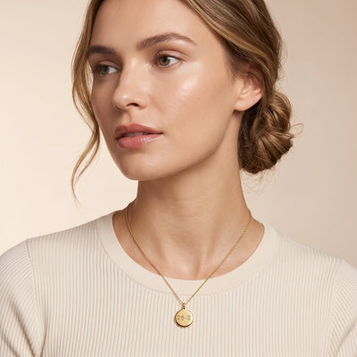A close-up of a woman wearing a delicate gold chain necklace with a round engraved gold pendant, styled with a cream ribbed knit top against a soft beige background.
