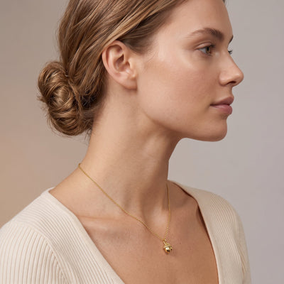 A woman in profile wearing a delicate gold chain necklace with a small gold acorn pendant, paired with a cream ribbed knit top against a soft beige background.
