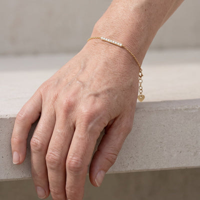 A close-up of a person's hand resting on a stone ledge, wearing a delicate gold chain bracelet with a short row of clear stones and a small dangling charm.