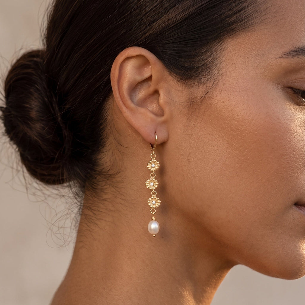A close-up side profile of a woman wearing a delicate gold dangle earring with a small floral cluster and a white pearl drop, set against a soft neutral background.