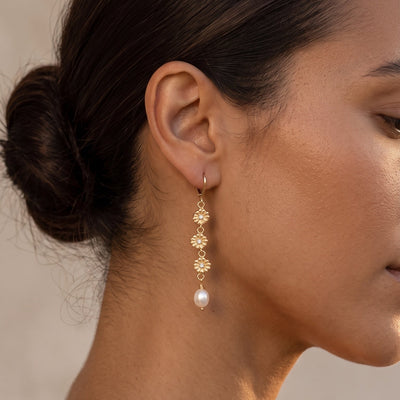 A close-up side profile of a woman wearing a delicate gold dangle earring with a small floral cluster and a white pearl drop, set against a soft neutral background.