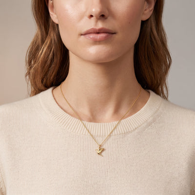 A close-up of a woman wearing a delicate gold necklace with a small bird pendant, styled with a cream-colored ribbed knit sweater against a soft neutral background.
