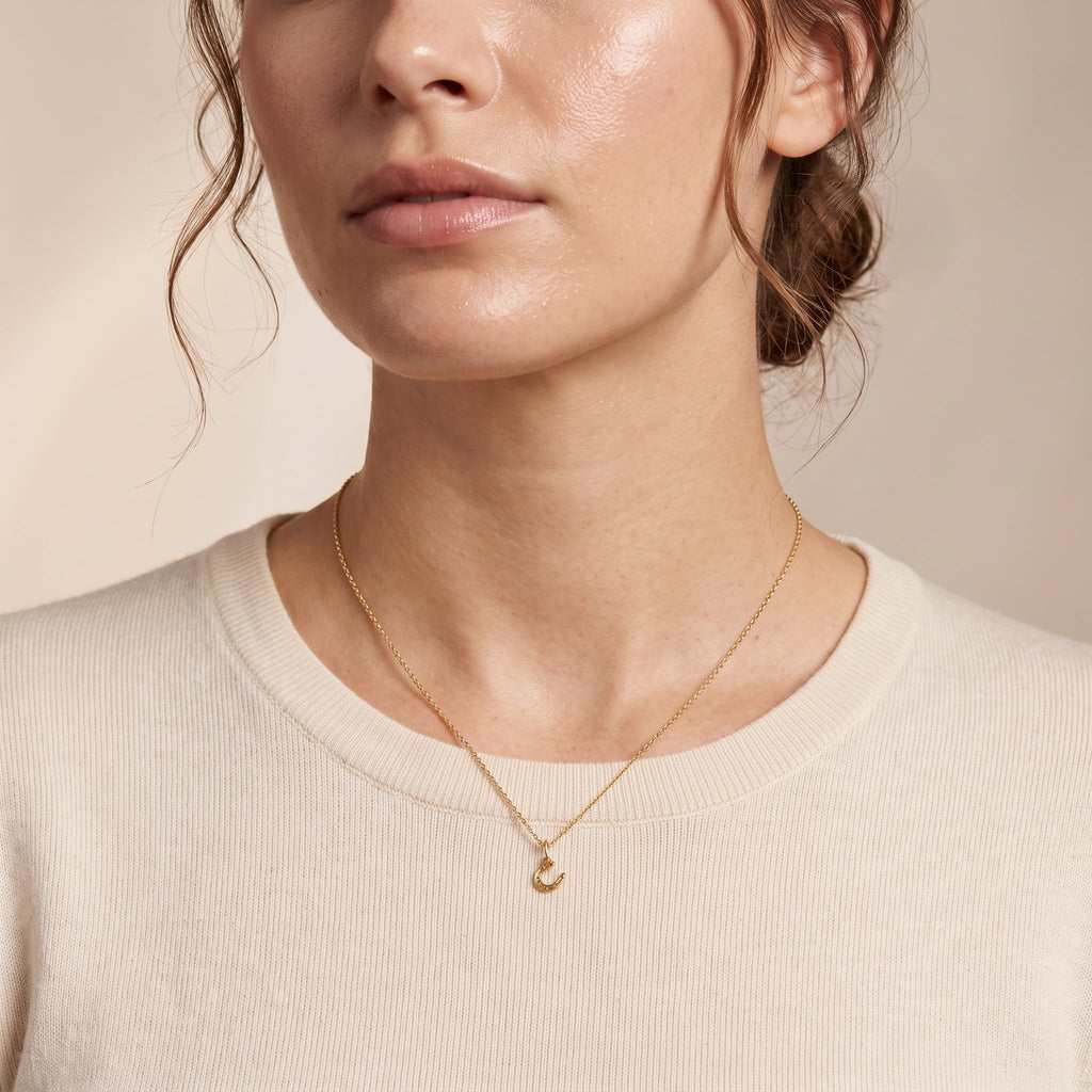 A close-up of a woman wearing a delicate gold necklace with a small crescent moon pendant, paired with a cream-colored crewneck sweater against a soft beige background.