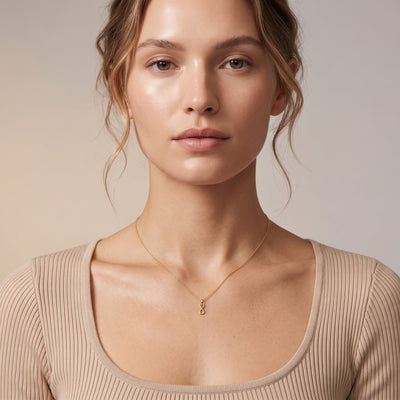 A close-up of a woman wearing a delicate gold necklace with a small pendant, paired with a beige ribbed scoop-neck top against a soft beige background.