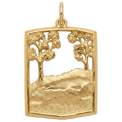 A gold pendant featuring an openwork landscape design with two stylized trees, a rocky hillside, and a fish motif in the center, shown on a plain white background.