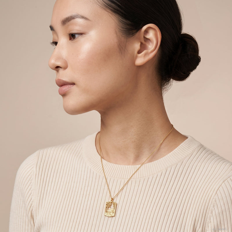 A close-up side profile of a woman wearing a delicate gold necklace with a rectangular pendant engraved with a landscape, paired with a cream ribbed knit top against a soft beige background.