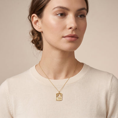 A close-up of a woman wearing a delicate gold pendant necklace with a rectangular landscape charm, styled with a cream-colored crewneck sweater against a soft beige background.