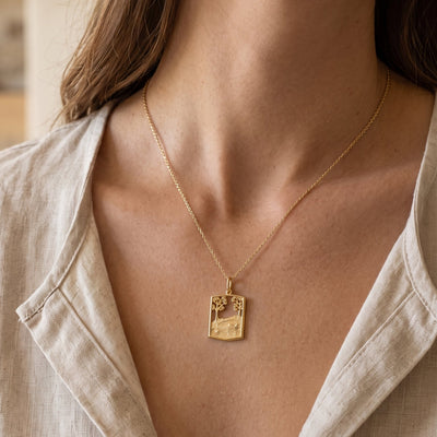 A close-up of a woman wearing a delicate gold chain necklace with a small rectangular pendant featuring a landscape design, paired with a light beige linen shirt.