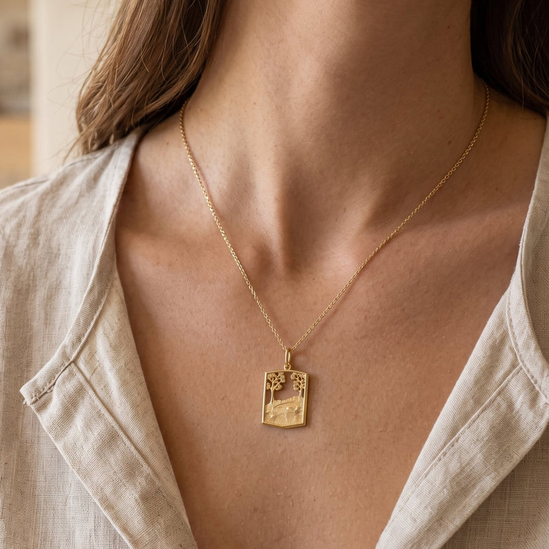 A close-up of a woman wearing a delicate gold chain necklace with a small rectangular pendant featuring a landscape design, paired with a light beige linen shirt.