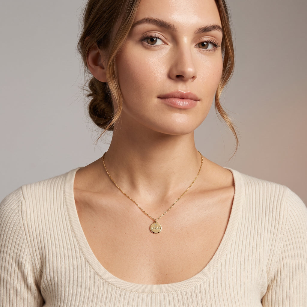A woman wearing a delicate gold chain necklace with a small round gold Mama pendant, paired with a cream-colored ribbed scoop-neck top, set against a neutral gray studio background.