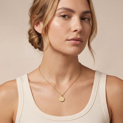 A woman wearing a delicate gold chain necklace with a small round gold pendant featuring an engraved Mimi, paired with a cream ribbed tank top against a warm beige background.