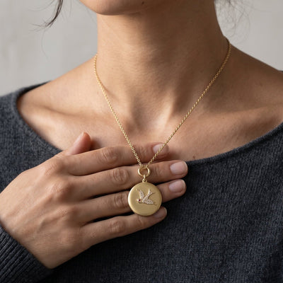 A close-up of a woman wearing a delicate gold chain necklace with a round, flat gold pendant featuring a bird design, set against a soft gray background.