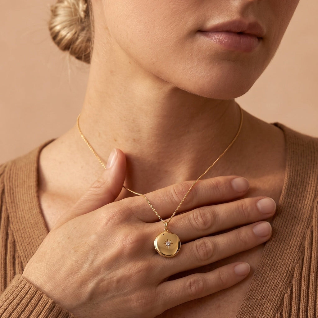 A close-up lifestyle shot of a woman wearing a delicate gold chain necklace with a small round pendant featuring a sunburst design, set against a warm beige background.