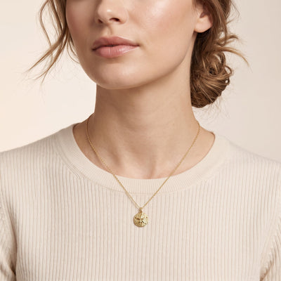 A close-up of a woman wearing a delicate gold necklace with a small, textured circular pendant, paired with a ribbed cream-colored top against a soft beige background.