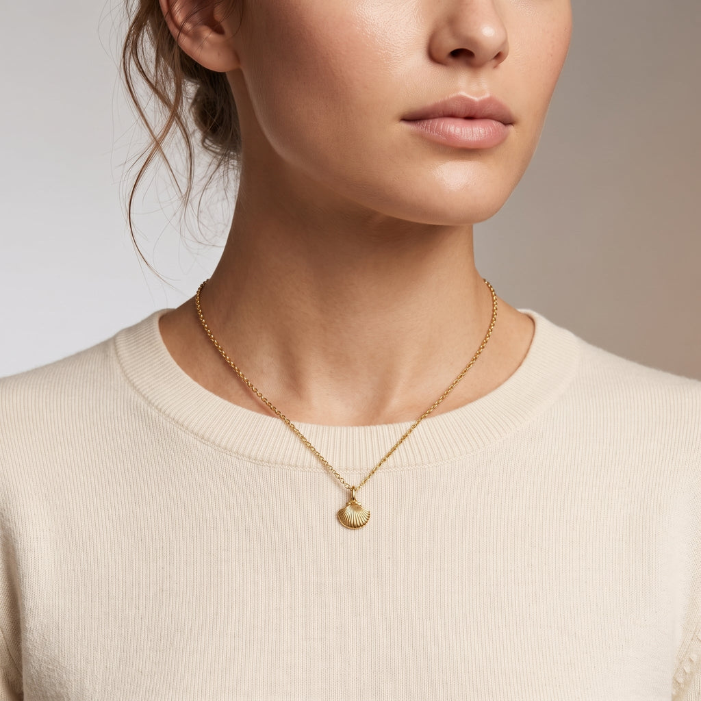 A close-up of a model wearing a delicate gold chain necklace with a small shell-shaped pendant, paired with a cream-colored crewneck sweater.