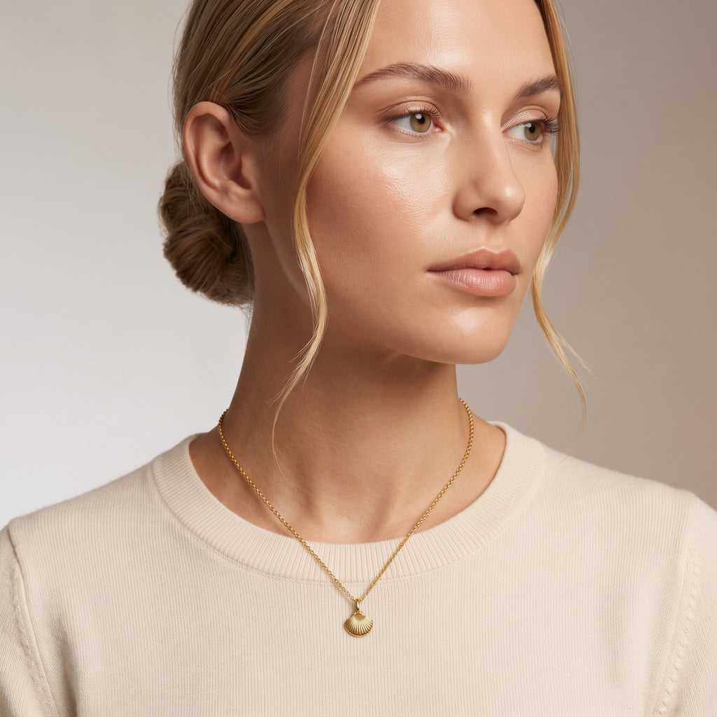 A close-up of a woman wearing a delicate gold chain necklace with a small scallop shell pendant, paired with a cream-colored crewneck sweater against a neutral beige background.