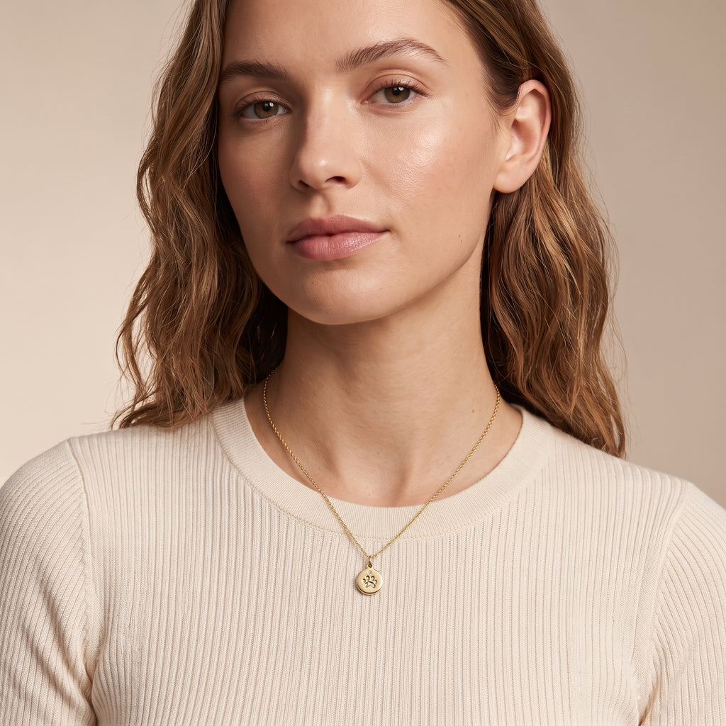 A close-up of a woman wearing a delicate gold necklace with a small circular pendant, paired with a cream-colored ribbed knit top, set against a warm beige background.