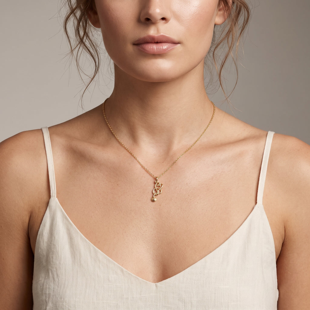 A close-up of a model wearing a delicate gold necklace with a small, stylized pendant, paired with a light beige linen camisole against a neutral gray background.