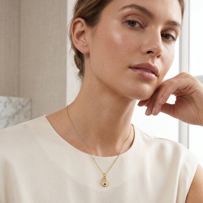 Woman wearing a gold necklace with a small pendant indoors.